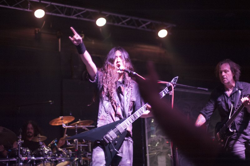 Masaki Murashita singing with one arm raised, accompanied by a guitarist and drummer under purple stage lights