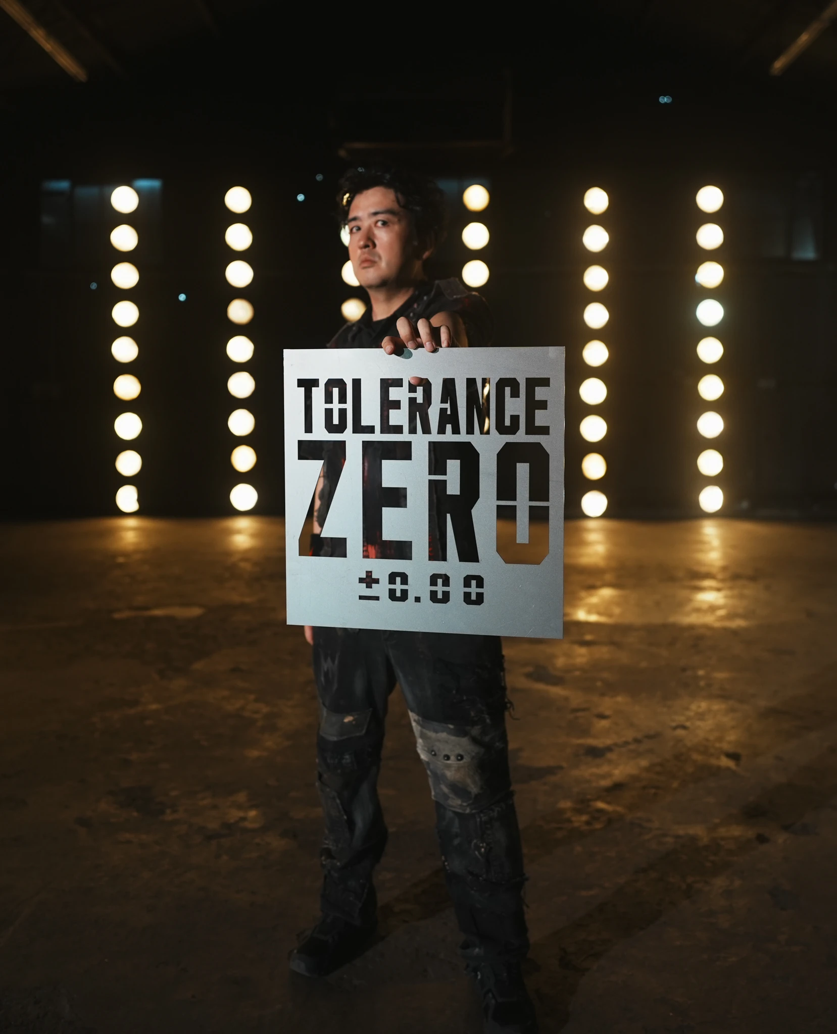 Masaki Murashita holding a cut metal Tolerance Zero plate in front of stage lights